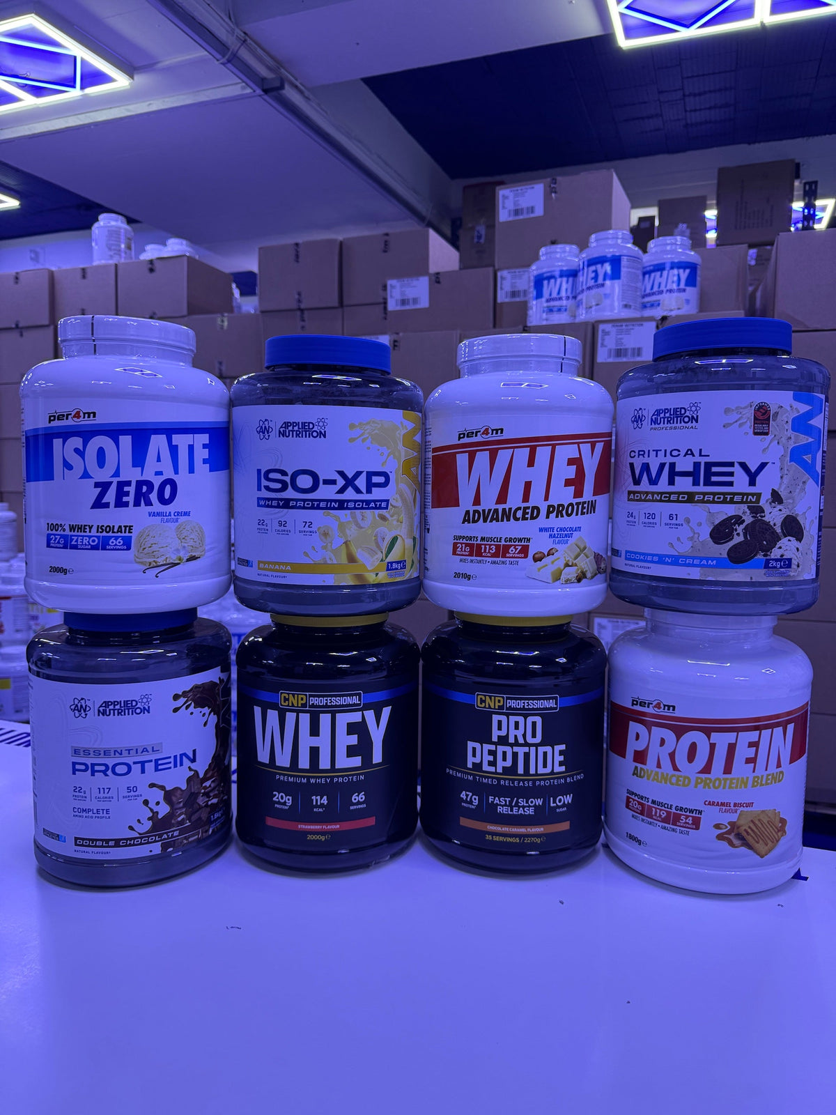 T-Nutrition warehouse shelf with Per4m, Applied Nutrition and CNP Professional whey protein tubs stacked together
